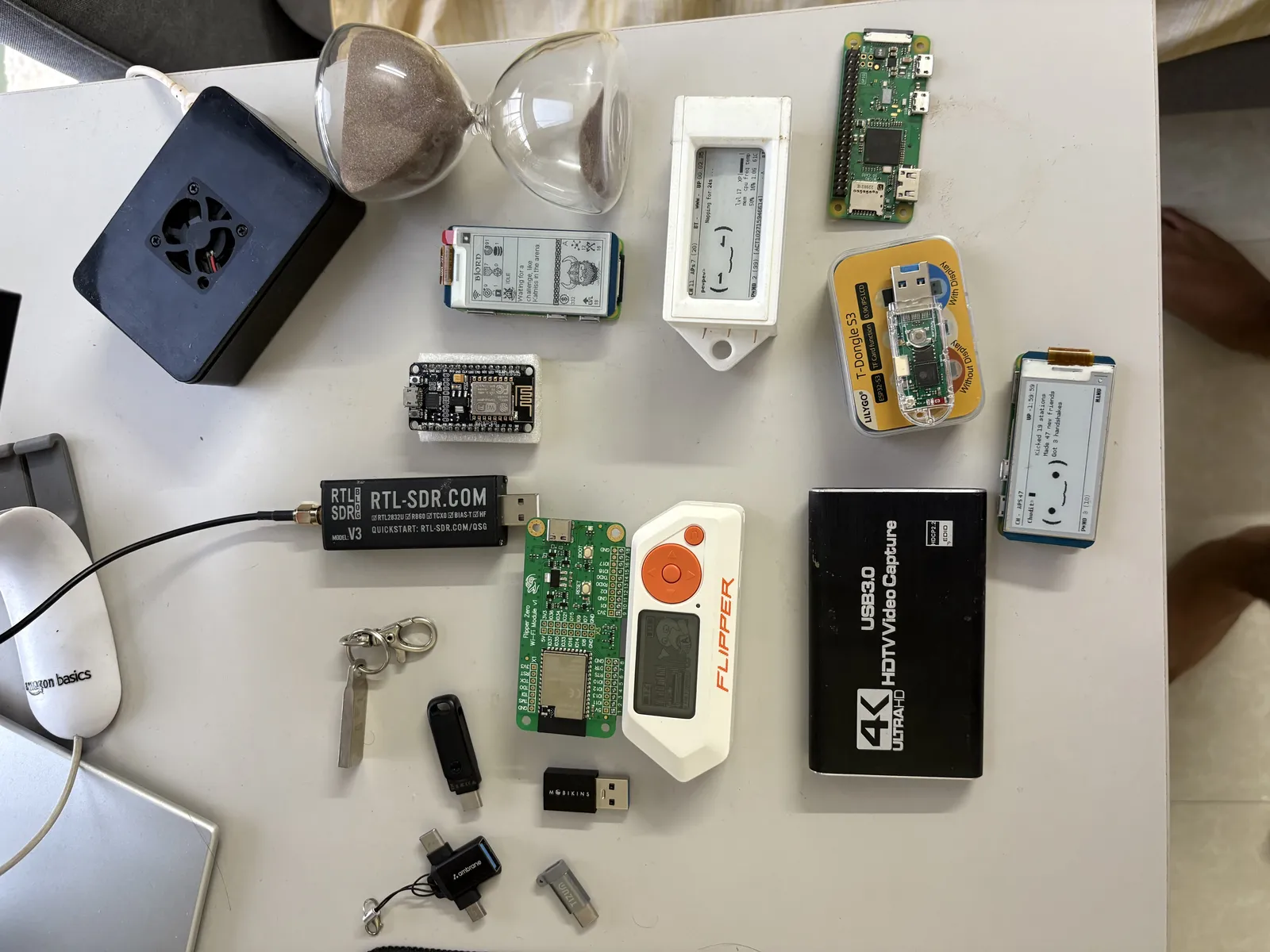 The workbench: SDR, Flipper Zero, Pwnagotchi, a Pi, and an hourglass for timing deploys.